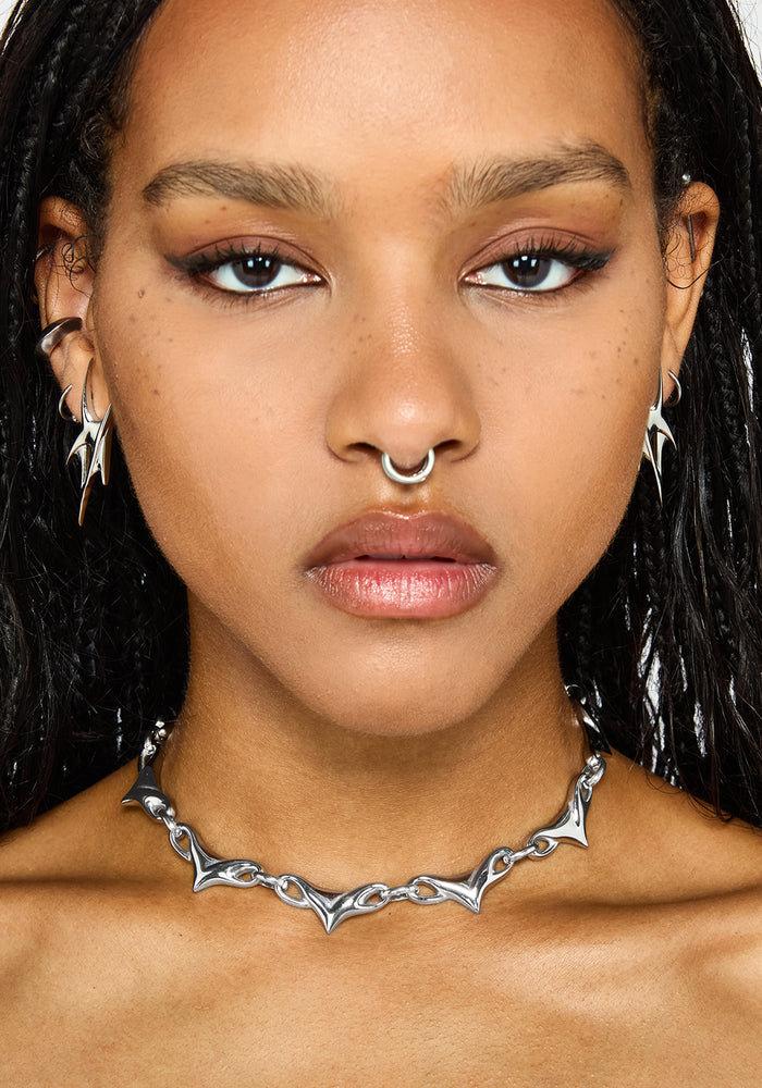 A close-up portrait of a model wearing a silver choker necklace with a spike, jagged, bat-wing-like design. The model also has other silver piercings and hoop earrings.
