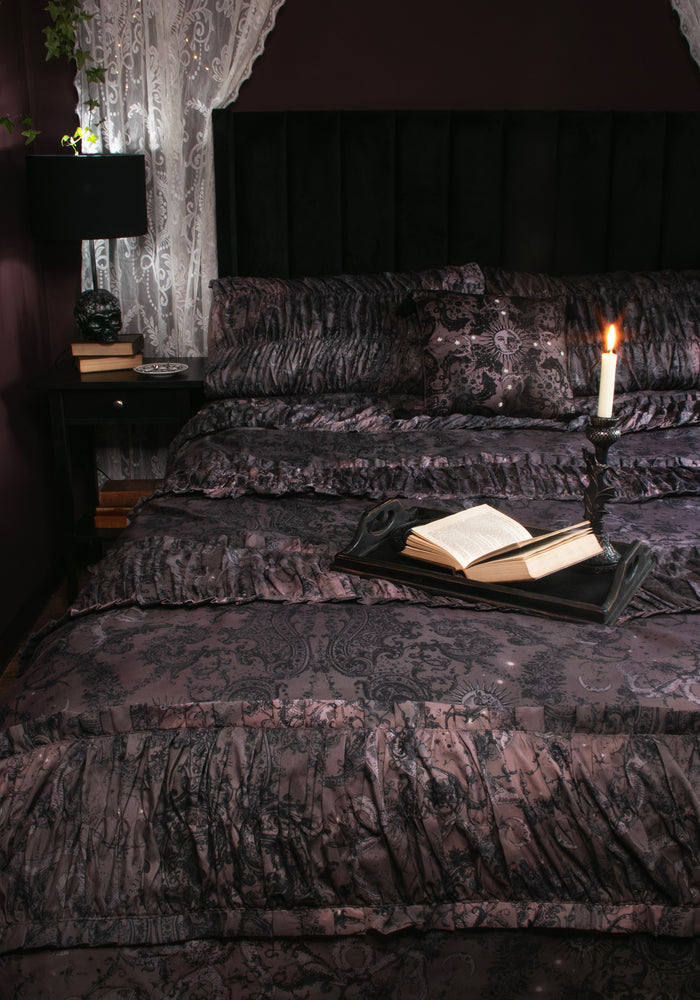 A dark, gothic-style bed dressed in black and deep purple damask-patterned bedding, featuring a black headboard, sheer lace curtains, a bedside lamp, and an open book on a tray with a lit candle.