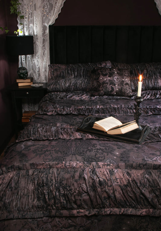 A dark, gothic-style bed dressed in black and deep purple damask-patterned bedding, featuring a black headboard, sheer lace curtains, a bedside lamp, and an open book on a tray with a lit candle.