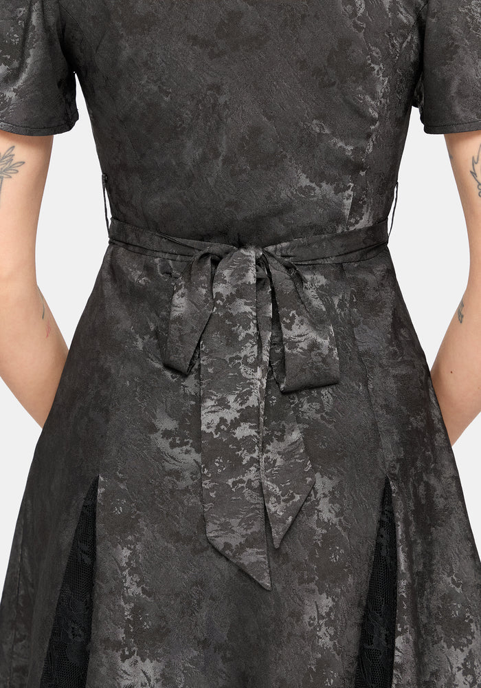 Close-up of a person wearing a dark charcoal grey short-sleeve mini dress with a textured jacquard pattern and a matching waist tie belt, shown from the back against a light gray background.