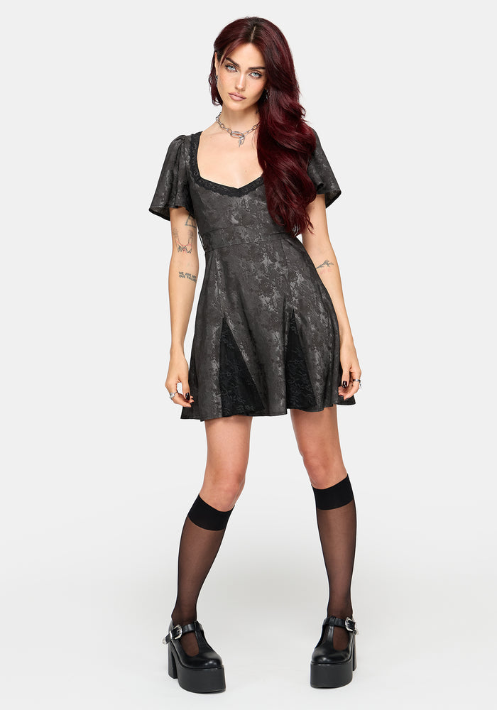 A model with long red hair stands against a light gray studio background wearing a short grey jacquard mini dress with sheer flutter sleeves, black thigh-high socks, and black platform Mary Jane shoes.