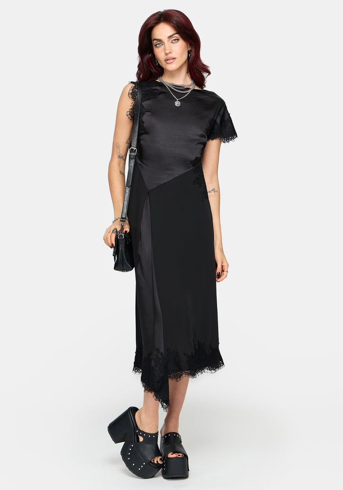 A woman modeling a black midi dress with sheer mesh side panels and tattered lace trim, styled with layered silver necklaces, a black shoulder bag, and black platform sandals against a light gray studio background.