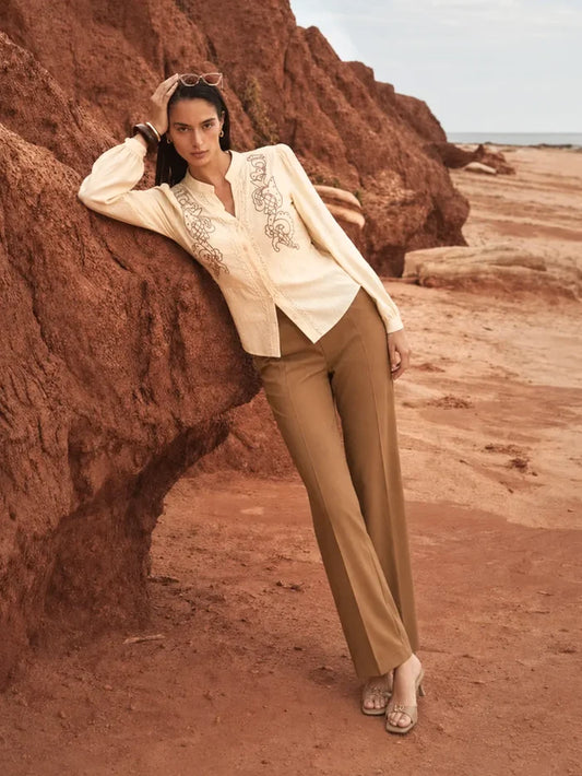 A woman posing outdoors against a large reddish-brown rock formation on a sandy beach, wearing a cream embroidered blazer, tan trousers, and metallic sandals.