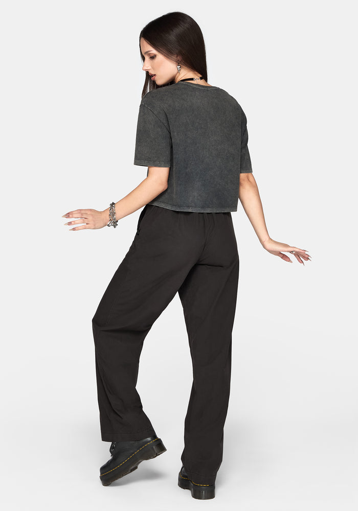 A woman posing from the back wearing a dark grey cropped t-shirt and black wide-leg trousers against a light gray background.