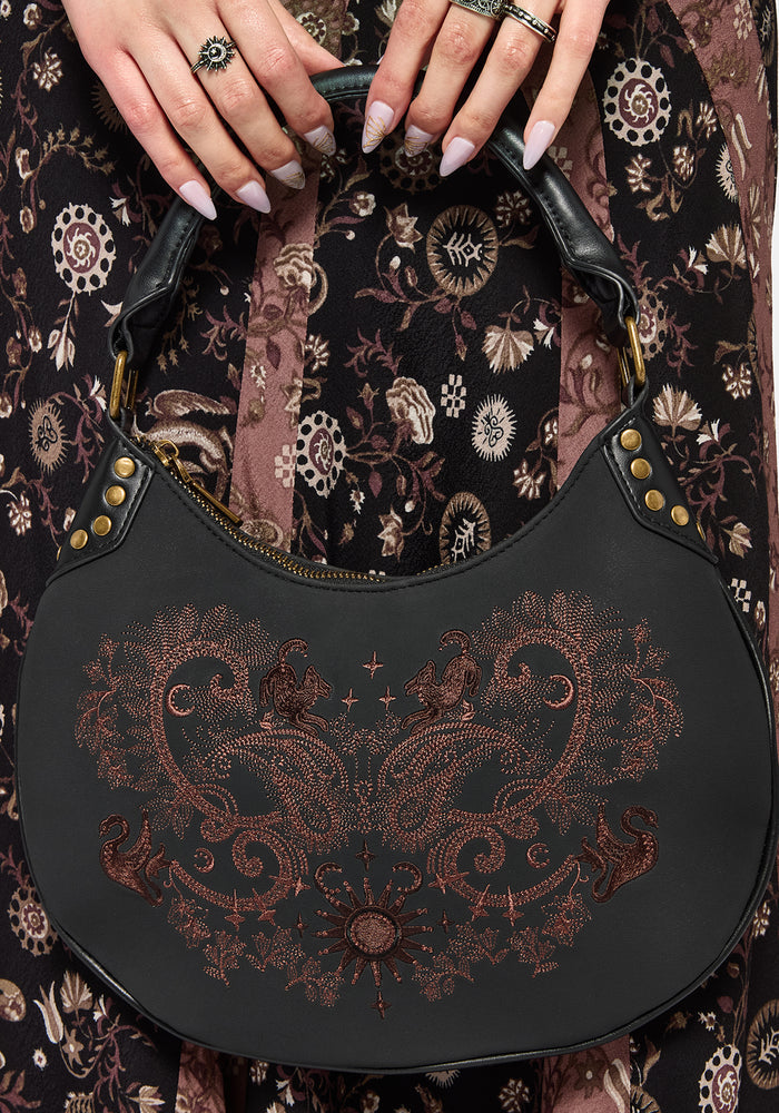 Black handbag with intricate brown embroidery held by a person against a patterned fabric background