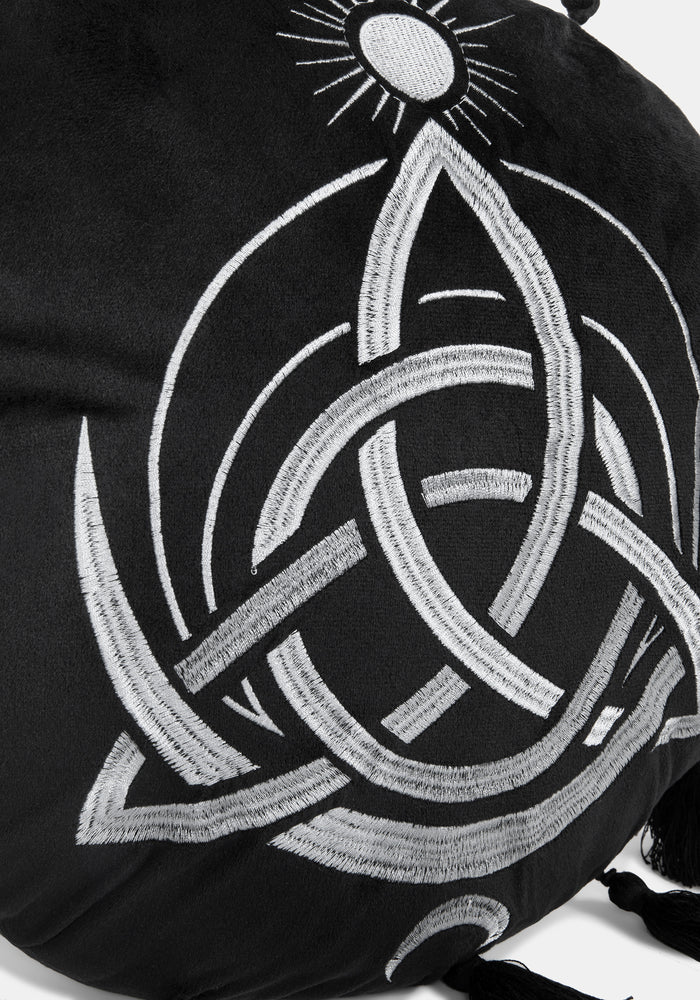 A circular black cushion with a white Celtic knot triquetra design, a small sun motif at the top, and tassels extending from the outer edge, shown on a light gray background.